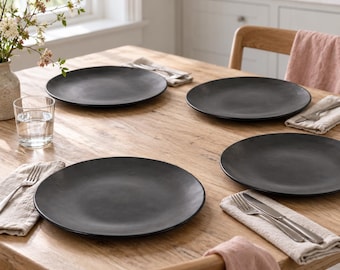 May include: A set of four round matte black dinner plates arranged on a wooden table. Each place setting includes a folded beige linen napkin, a silver fork, and a silver knife. A clear glass of water sits near a vase with small white flowers.