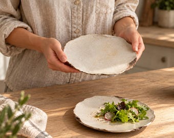 May include: Two round, off-white ceramic plates with irregular, organic edges and a speckled, matte texture. One plate is held up, while the other rests on a surface, containing a fresh green salad with sliced radishes.