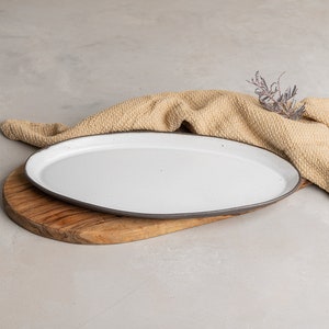 May include: An oval ceramic serving platter with a smooth white glazed surface and a dark grey rim. The platter rests on a natural wooden board with a visible grain pattern, accompanied by a textured tan fabric cloth and a small dried plant sprig.