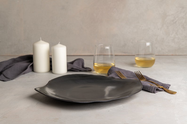May include: A dark gray, irregularly shaped ceramic plate with a smooth, reflective surface. The plate features a wide, organic rim and a shallow center, presented alongside two gold-toned metal forks, two white pillar candles, and two glasses.