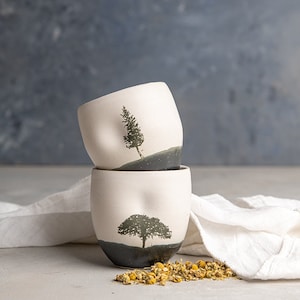 May include: Two off-white ceramic cups stacked vertically, each featuring a dark gray base and a black silhouette of a tree. The top cup displays a tall pine tree, while the bottom cup shows a wide, rounded tree. The surface of the cups is matte.