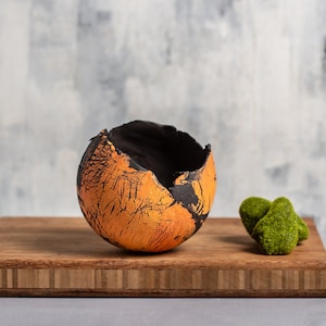 May include: A small, orange and black ceramic planter with a cracked glaze finish. The planter is sitting on a wooden surface.