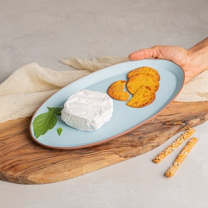 May include: A blue oval ceramic serving platter with a round white cheese, green basil leaves, and several round crackers on a wooden chopping board.
