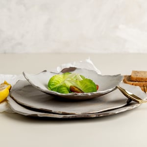 Könnte beinhalten: Ein weißer Keramikteller mit einem Salat aus Salat und Tomaten darauf. Der Teller steht auf einem Tisch mit einer weißen Tischdecke und einer Zitronenscheibe daneben. Auf dem Tisch liegen zwei goldene Gabeln und ein goldener Löffel.