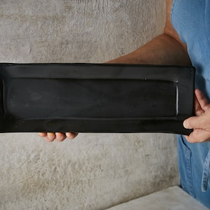 Könnte beinhalten: Eine rechteckige Servierplatte mit einer glatten, mattschwarzen Oberfläche. Das Keramiktablett verfügt über leicht erhöhte Ränder und eine flache, längliche Form, die ein schlichtes Design für die Präsentation verschiedener Speisen bietet.