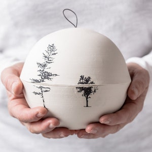 May include: A white ceramic urn with a lid, featuring a black and white tree design. The urn is being held in the hands of a person.