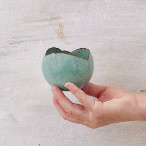 May include: A small, teal-colored ceramic bowl with an uneven, slightly chipped rim. The bowl is held in a hand against a white wood background.