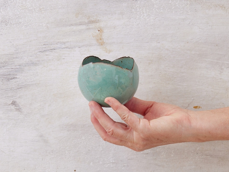 May include: A small, teal-colored ceramic bowl with an uneven, slightly chipped rim. The bowl is held in a hand against a white wood background.