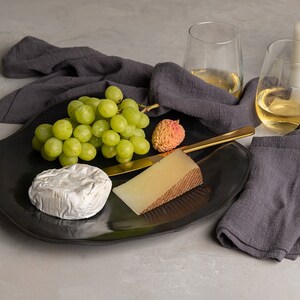 May include: A black, irregularly shaped serving platter holds a cluster of green grapes, a single lychee, a round soft-ripened cheese, a wedge of firm cheese with a brown rind, and a gold-toned cheese knife. Two glasses of white wine and two white candles.