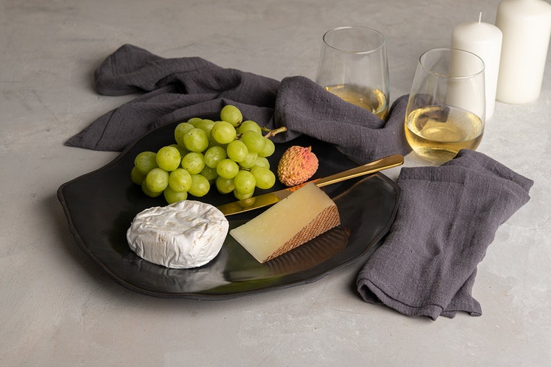 May include: A black, irregularly shaped serving platter holds a cluster of green grapes, a single lychee, a round soft-ripened cheese, a wedge of firm cheese with a brown rind, and a gold-toned cheese knife. Two glasses of white wine and two white candles.