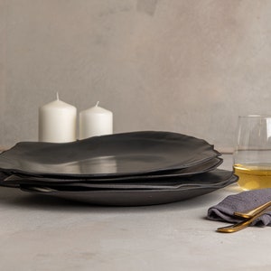 May include: A stack of three matte black ceramic plates with irregular, organic edges. The plates are positioned near two white pillar candles, two clear glasses filled with a light-colored liquid, a dark gray cloth napkin, and a gold-toned fork.