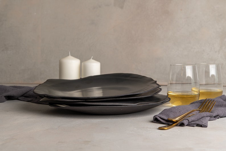 May include: A stack of three matte black ceramic plates with irregular, organic edges. The plates are positioned near two white pillar candles, two clear glasses filled with a light-colored liquid, a dark gray cloth napkin, and a gold-toned fork.