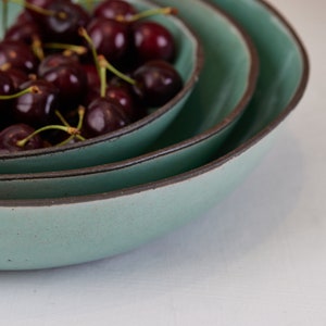 May include: A stack of three round ceramic bowls with a matte sage green interior and a dark brown rim. The smallest bowl at the top is filled with a cluster of fresh, dark red cherries with green stems, showcasing a speckled texture.