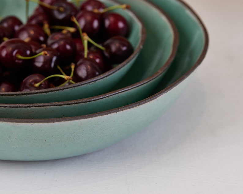 May include: A stack of three round ceramic bowls with a matte sage green interior and a dark brown rim. The smallest bowl at the top is filled with a cluster of fresh, dark red cherries with green stems, showcasing a speckled texture.