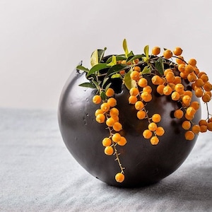May include: A dark, round ceramic vase with a textured surface. The vase is filled with green leaves and cascading strands of small, bright orange berries. The arrangement sits on a light gray surface.