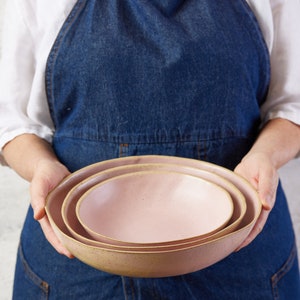 May include: A set of three nested ceramic bowls in a soft pastel pink hue. Each bowl features a smooth, rounded interior and a slightly speckled, textured exterior finish with a natural, earthy rim that contrasts with the light pink glaze.