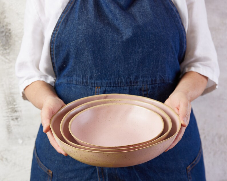 May include: A set of three nested ceramic bowls in a soft pastel pink hue. Each bowl features a smooth, rounded interior and a slightly speckled, textured exterior finish with a natural, earthy rim that contrasts with the light pink glaze.
