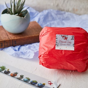 May include: A red tissue paper package with a white label that reads "Dannah Levy Ceramics Artist" and includes contact information. The package is sitting on a white surface with a blue fabric in the background.