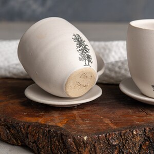 以下が含まれることがあります： 黒い松の木のグラフィックが施された、オフホワイトの陶器製マグカップ2個とソーサーのセット。片方のマグカップは横向きに置かれ、釉薬のかかっていない底面にClay by Pamelaという文字が刻まれており、もう片方はソーサーの上に直立しています。