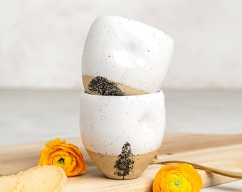 May include: Two stacked ceramic cups with a speckled white glaze on the upper portion and a natural tan base. Each cup features a small thumb indentation and a black printed tree silhouette. The cups are accompanied by yellow flowers and gold spoons.