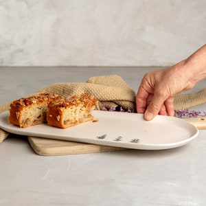 May include: A cream-colored, oval-shaped serving platter with a minimalist tree design. Two slices of cake with a nut topping sit on the platter. A hand is holding the platter, ready to serve.