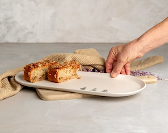 May include: An elongated, off-white ceramic serving platter with a smooth finish and small, dark, tree-like illustrations. Two slices of almond-topped cake sit on the left side of the platter, which features a handle on the right end.