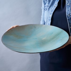 Op de afbeelding: Grote, ovale keramische serveerschaal met een lichtblauw en bruin aquarelontwerp. De schaal heeft een donkere rand en wordt vastgehouden door een persoon met een spijkerjasje. Dit decoratieve stuk is ideaal om eten te serveren of als decoratief accent.