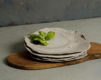 May include: A stack of three off-white ceramic plates with irregular, organic edges. The plates have a smooth, matte finish and are resting on a long, oval-shaped wooden board. A small cluster of fresh green lettuce leaves sits on the top plate.