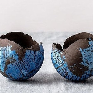 May include: Two ceramic bowls with a cracked, eggshell-like design. The bowls are primarily blue with dark brown accents around the rim. The interior of the bowls is dark brown. The bowls are on a light gray surface.