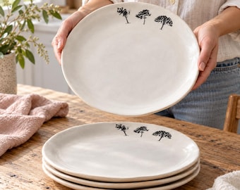 May include: A set of off-white ceramic plates with an uneven, organic edge. Each plate features a small, black, hand-drawn illustration of three distinct trees positioned near the upper rim of the circular dinnerware.