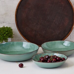 May include: Three round, shallow bowls with a glossy, light mint-green interior and a dark brown rim. The bowls have an organic, slightly irregular shape. One smaller bowl is filled with dark red cherries, and one single cherry sits nearby.