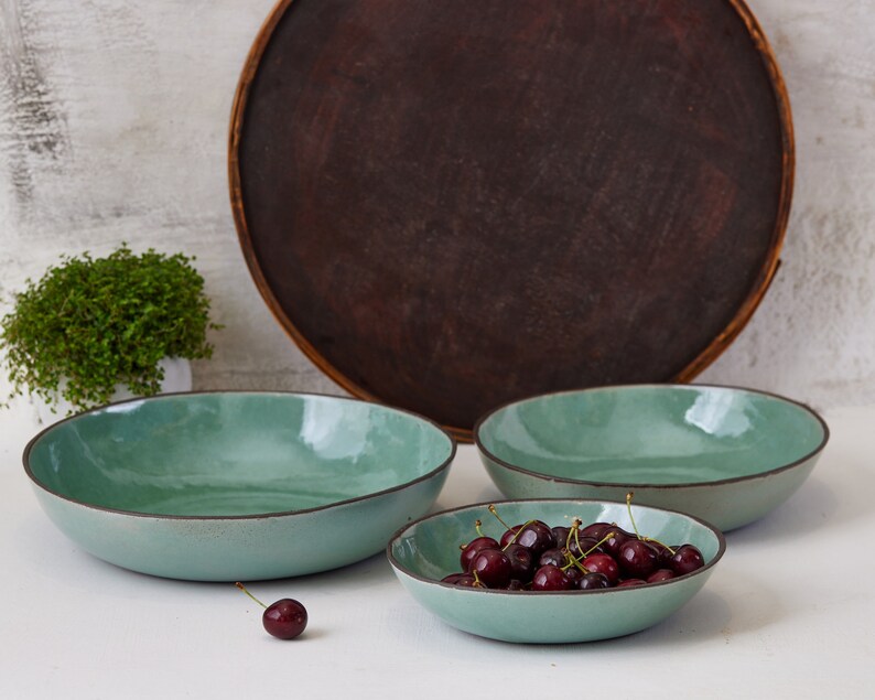 May include: Three round, shallow bowls with a glossy, light mint-green interior and a dark brown rim. The bowls have an organic, slightly irregular shape. One smaller bowl is filled with dark red cherries, and one single cherry sits nearby.