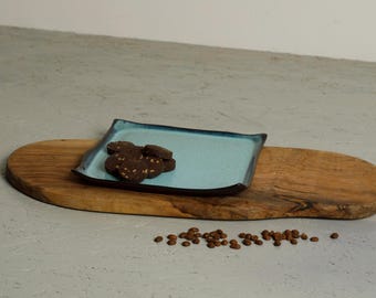 May include: A square ceramic plate with a light blue center and dark blue edges, holding a small pile of round dark chocolate cookies. The plate rests on an elongated, oval-shaped wooden board with a natural grain, accompanied by scattered coffee beans.