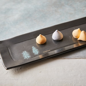 May include: A black rectangular serving tray with a light blue tree design and the text "Clay by Design". Four meringue cookies are arranged on the tray: two yellow, one purple, and one white.