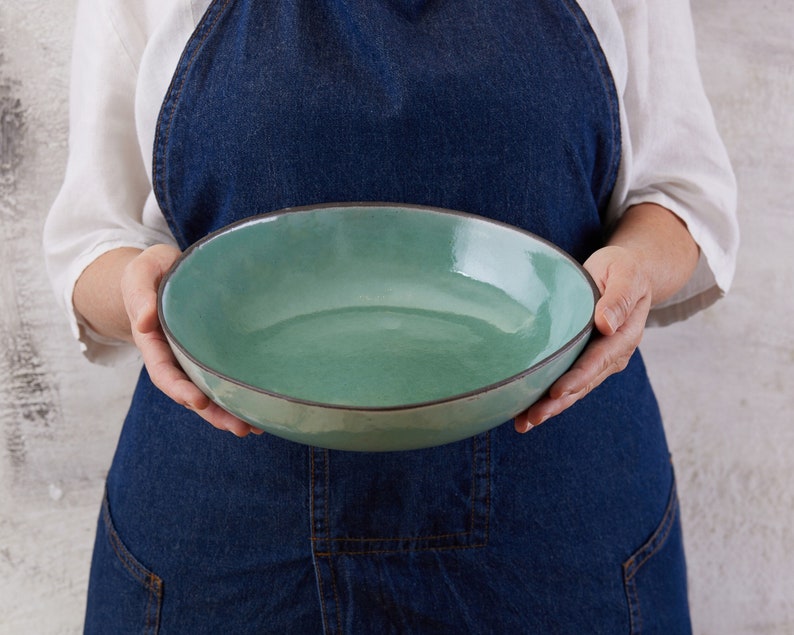 May include: A round ceramic bowl with a glossy sage green interior and a dark brown rim. The bowl features a smooth, slightly irregular edge and a shallow basin, showcasing a handcrafted appearance with a reflective surface finish.