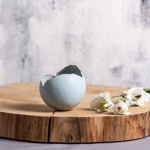 May include: A light blue ceramic egg-shaped vase with a crack on the top, sitting on a wooden slice with white flowers.