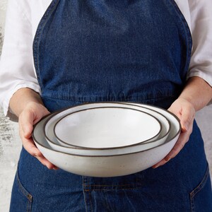 May include: A set of two nested round bowls with a smooth white interior and a dark brown rim. The bowls feature a speckled texture on the exterior surface and a shallow, wide basin design, crafted from ceramic material with a clean, simple finish.