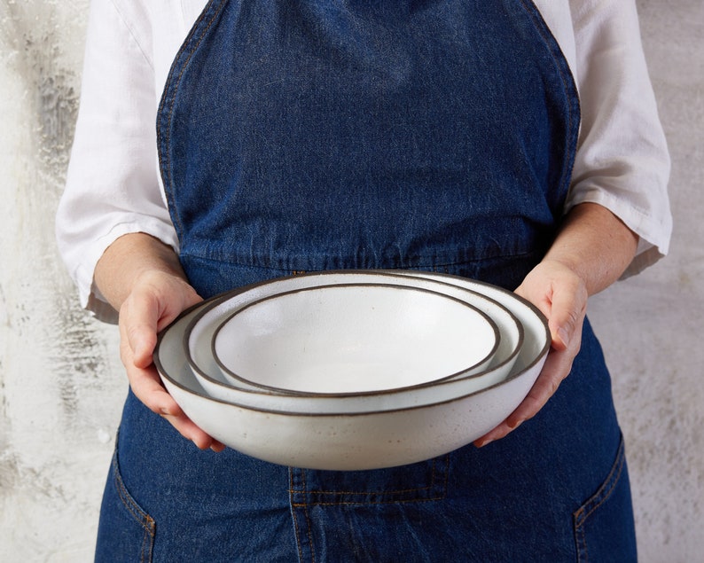 May include: A set of two nested round bowls with a smooth white interior and a dark brown rim. The bowls feature a speckled texture on the exterior surface and a shallow, wide basin design, crafted from ceramic material with a clean, simple finish.