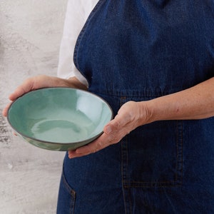May include: A round ceramic bowl with a glossy, light turquoise interior and a dark, matte charcoal exterior rim. The bowl features a smooth, slightly irregular edge and a reflective surface that catches the light, showcasing a simple, functional design.
