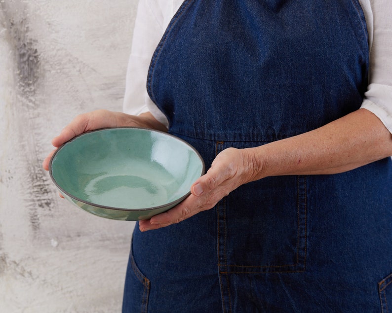 May include: A round ceramic bowl with a glossy, light turquoise interior and a dark, matte charcoal exterior rim. The bowl features a smooth, slightly irregular edge and a reflective surface that catches the light, showcasing a simple, functional design.