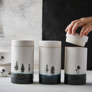 May include: Three white ceramic canisters with black tree silhouettes and a dark blue glaze at the bottom. The canisters have lids and are arranged in a row on a white surface.