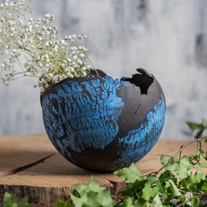 May include: A blue and black ceramic vase with a crackled finish. The vase is round and has a chipped edge. It is filled with white flowers and sits on a wooden surface.