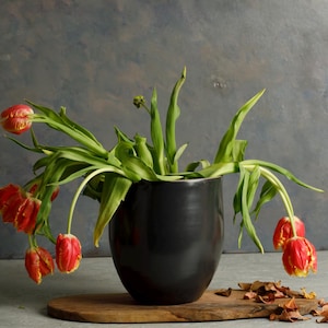 May include: A black ceramic vase with a wide mouth holds a bouquet of wilting red and yellow tulips. The vase is sitting on a wooden surface.
