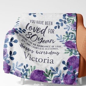 May include: A white throw blanket with a floral design in shades of purple and blue. The text "You have been loved for 80 years Wishing you an abundance of love, joy, and countless blessings. Happy birthday Victoria" is printed on the blanket.