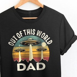 May include: Black t-shirt with a retro-style graphic of three flying saucers and the text "Out of this World Dad".