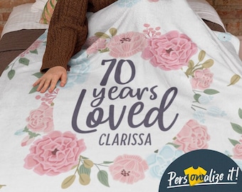 May include: A white floral blanket with the text "70 years loved Clarissa" in gray lettering. The blanket features pink and blue flowers.