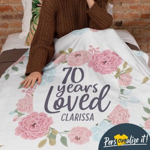 May include: A white floral blanket with the text "70 years loved Clarissa" in gray lettering. The blanket features pink and blue flowers.
