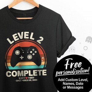 May include: Black t-shirt with a retro-style video game controller graphic and the text "Level 2 Complete". The graphic has a sunset color scheme and the text is in white. The shirt also includes the text "Scott ♥ Mary - Since March 18, 1988 - "