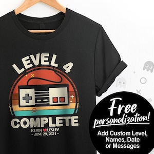 May include: Black t-shirt with a retro-style graphic of a video game controller. The graphic says "Level 4 Complete" with the text "Kevin ♥ Lesley - June 29, 2021 -" below the controller.