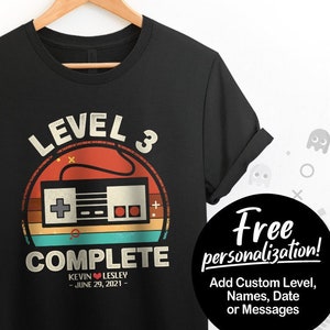 May include: Black t-shirt with a retro video game controller graphic. The design includes the words "LEVEL 3 COMPLETE" and the names "KEVIN LESLEY" with a date. The shirt is hanging on a wooden hanger.